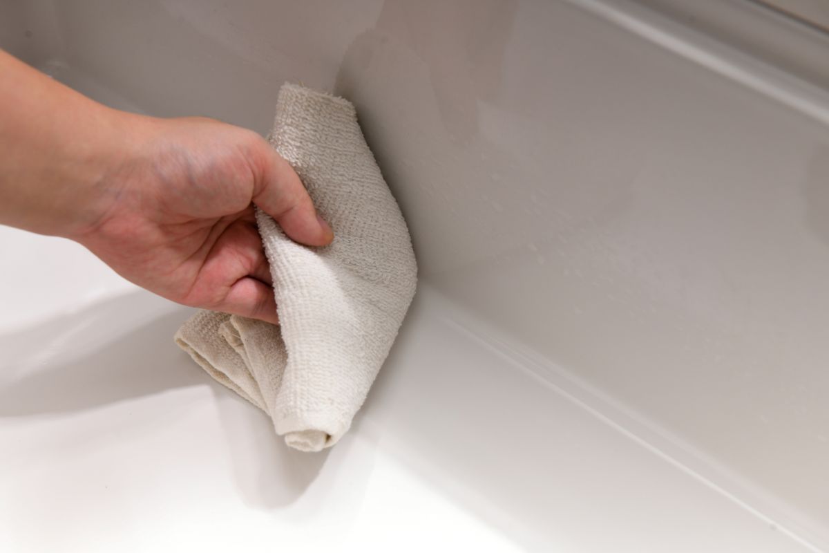 A hand wiping the glazed interior of a white fireclay kitchen sink with a soft cloth — the mild daily-care routine the fired, non-porous surface asks for